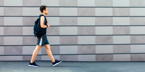 Fototapeta premium Go to morning workout. Guy in sportswear, sneakers and with backpack, on gray wall background, side view, free space