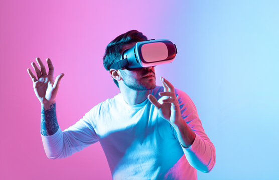 A man interacts with virtual reality technology, wearing a VR headset while standing against a colorful backdrop. His hands are raised as he explores immersive digital environments.