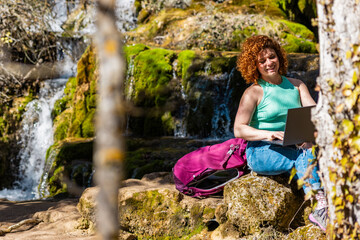 Freelancer working remotely on laptop near waterfall during hiking trip