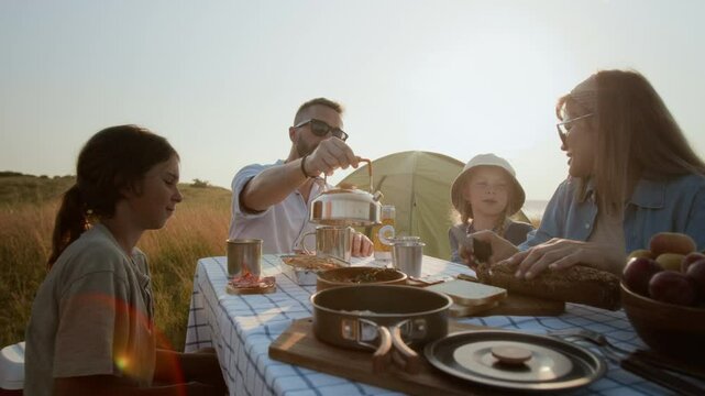 Family sitting at picnic table during camping trip, father pouring tea for kids and mother slicing bread during golden hour on beautiful lakeside