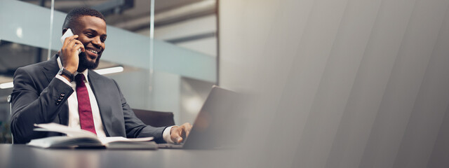 Successful young black businessman in expensive blue suit working on laptop at office, having phone conversation with his personal assistant or business partner and smiling, copy space