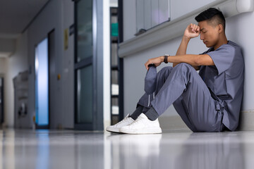 Sitting in hospital corridor, healthcare worker in scrubs looking thoughtful and tired, copy space