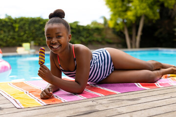 Smiling girl in striped swimsuit enjoying popsicle by pool on sunny day, copy space
