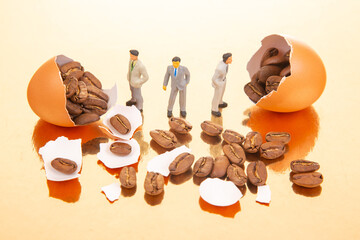 Small figurines in professional and casual outfits stand by orange eggs bursting with coffee beans on a shiny golden surface, symbolizing the fusion of business and natural elements.