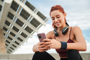 Happy young woman enjoying smartphone while sitting outdoors with headphones, engaged in mobile...