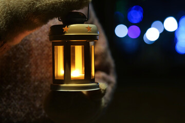 Woman holding Christmas lantern with burning candle in darkness, closeup and space for text. Bokeh effect