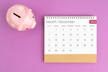 December 2025 desk calendar and pink piggy bank on purple background.