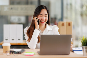 Busy young asian business woman, young professional lady executive manager talking on the phone making business call on cellphone sitting at work desk in office using laptop computer.
