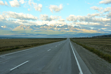 An asphalt road crosses the morning steppe in the shadow of clouds to meet the snow-capped mountain peaks in late summer.