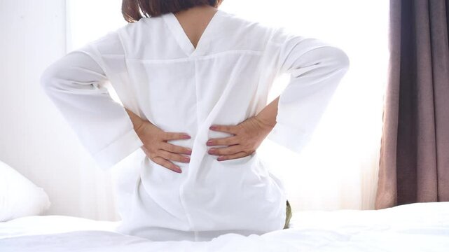 Asian woman waking up in bed with lower back pain due to bad sleeping position, feeling stiff and uncomfortable in the morning
