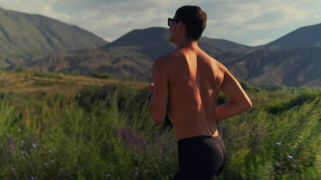 A sun kissed runner topless in athletic shorts and sunglasses runs along a road through green slopes. The camera captures him from the side as the setting sun highlights his muscular frame.