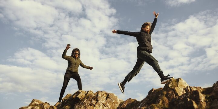 Two people jumping on rocky terrain under a cloudy sky. Outdoor adventure and fun on rocks. Energetic leap on rocks, enjoying nature and rocky landscape. Hiking adventure on mountain trail.