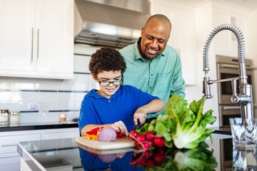 Father and son cooking together in a modern kitchen. Smiling African American father helps son chop vegetables. Family bonding in the kitchen. Black father and son cooking family lunch in kitchen.