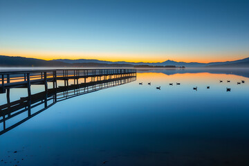 Obraz premium Tranquil Sunrise Over Serene Lakeside with Wooden Pier and Reflective Waters in Peaceful Natural Setting
