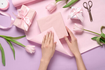 Young woman wrapping gift boxes with tulips on lilac background. International Women's Day