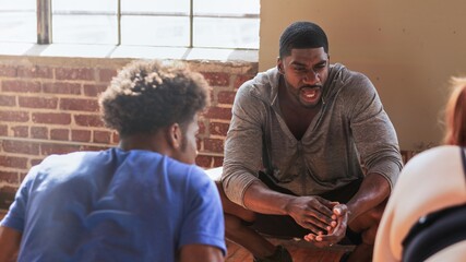 A group of people with coach, engaged in conversation. They are sitting near a window with brick...