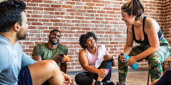 Group of diverse individuals in workout attire, smiling and chatting in a gym setting with a brick wall background, holding water bottles and towels. Diverse people talking after gym class.