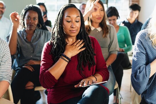 A diverse group of people in a workshop setting, focusing on mindfulness. Diverse are seated, with one woman in the foreground practicing a calming exercise with hand on her chest.
