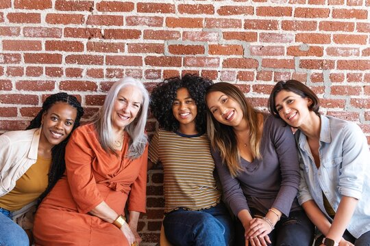 Group of five diverse women sitting together, smiling in front of a brick wall. Diverse ethnicities and different ages, casual attire, friendly atmosphere, cheerful expressions.