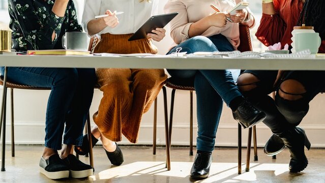 Diverse women sitting at a table, focused on work. Diverse women sitting in row, working on tablets and notebooks. Collaborative workspace and teamwork. Diverse women discussing and working together