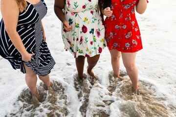 Curvy women in colorful dresses stand in ocean waves. Beach fun with floral and striped dresses. Summer dresses, ocean waves, and beach vibes. Curvy women at beach in summer wearing summer dresses.