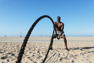 Man exercising with battle ropes on a sandy beach. Intense workout with ropes on the beach. Athletic man focused on rope training outdoors. African American man doing exercise workout photography.