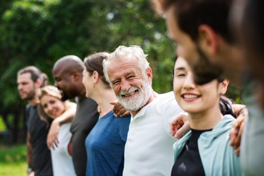 Elderly man and diverse people hugging each other. Diverse group of women and men embrace each other at park. Unity and teamwork. Backside of old man and friends standing in row huddle on green grass.