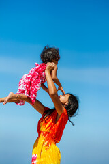 Asian teenage girl in colorful beach dress holding and lifting up her little sister, playing during summer vacation with blue sky on the background
