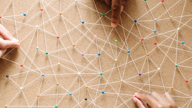 Hands connecting string on a corkboard, creating a network pattern. Diverse hands, teamwork, and collaboration. Diverse people connecting string as network pattern. Network string pattern connection.