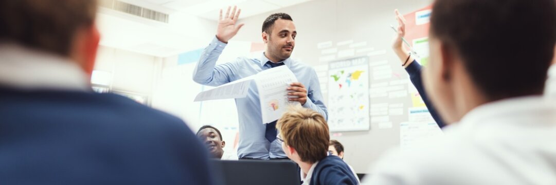 Man teacher engages students in a classroom, holding papers and raising a hand. Diverse students listen attentively, interactive learning. Man teacher in class hands up with students participate - Powered by Adobe
