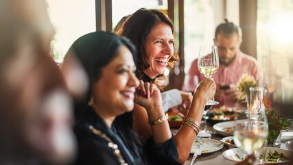 Diverse women smiling amd emjoying meal, holding wine glasses, lively atmosphere, dining together, cheerful gathering, celebrating with friends. Happy friends toasting and celebrating at restaurant.