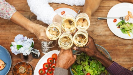 Diverse group of friends toasting with champagne glasses over a wooden table top view. Diverse people celebration with champagne in restaurant. Diverse friends celebrating together during lunch.
