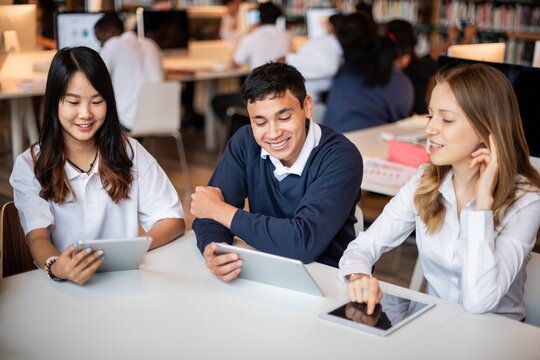 Students studying together in a library, using tablets. Diverse group of young adults collaborating and engaging with technology in an academic setting. Diverse high school students at library.