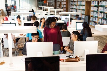 Teacher and diverse students studying in a library with computers. Diverse young adults studying and learning with computers. Students talk with teacher in library with computers. Learning in library