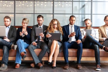 Group of diverse professionals sitting on a bench, using laptops and phones. Business women and...