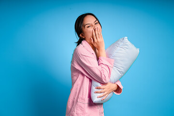 Asian woman in pink pajamas yawns while hugging a white pillow, showing signs of tiredness and...