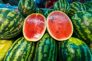 A pile of fresh whole watermelons with one cut open to reveal vibrant red flesh and dark seeds, displayed at a market stall
