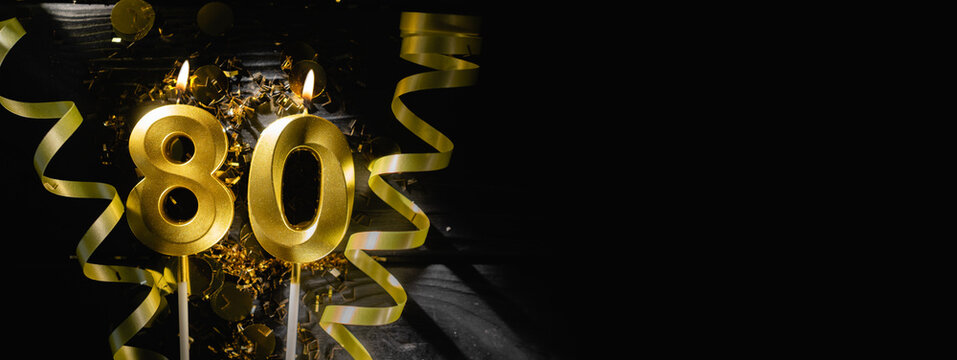 Golden candles number eighty burning brightly, celebrating 80th birthday anniversary. Festive ribbons and confetti on black background, creating elegant celebratory banner.