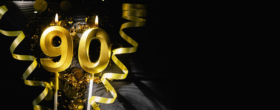 Golden candles number ninety burning brightly, celebrating 90th birthday anniversary. Festive ribbons and confetti on black background, creating elegant celebratory banner.