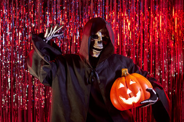 Portrait of the grim reaper holding a orange pumpkin on a red background during a halloween party....