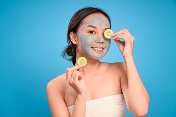 Asian woman with a gray clay mask on her face playfully holds cucumber slices near her eyes while...