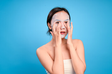 Fototapeta premium sian woman wearing a white sheet mask gently touches her cheeks with both hands, looking to the side with a calm expression, standing against a blue background, promoting skincare and beauty care