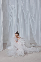 woman clad in a flowing white gown sits gracefully on the floor, surrounded by ethereal draped...