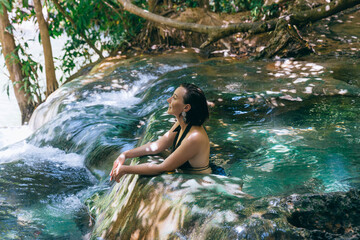 Fototapeta premium person enjoys a tranquil moment in a natural hot spring, sitting on a smooth rock with clear water cascading around. Sunlight filters through the trees, creating a serene atmosphere