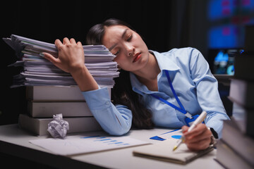 Young office worker resting her head on a large pile of paperwork, feeling overwhelmed and...