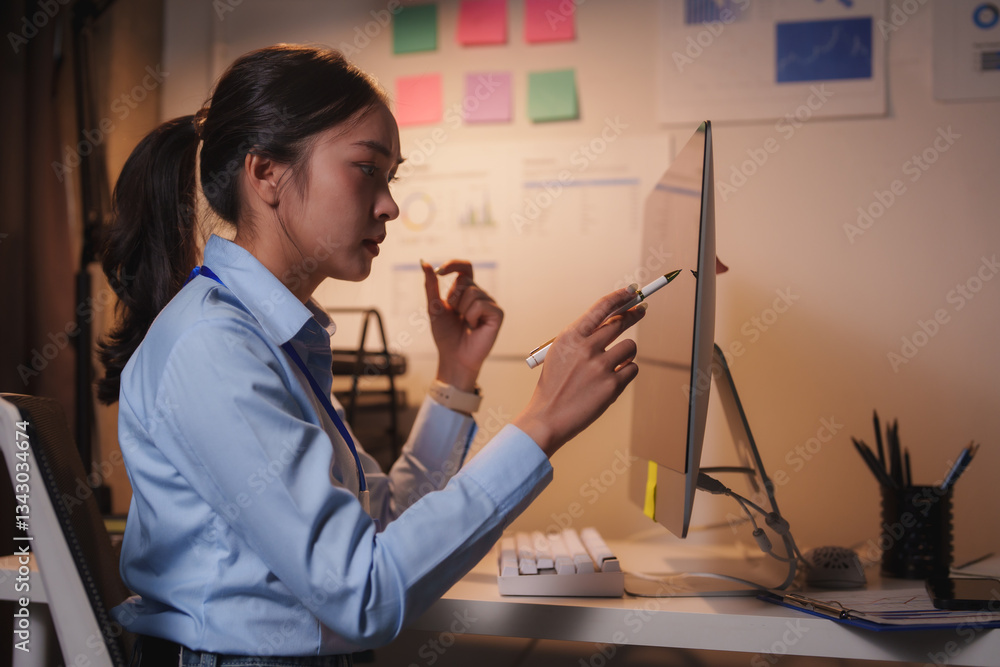 Poster young businesswoman pointing at computer monitor with a pen while working late in the office, analyz