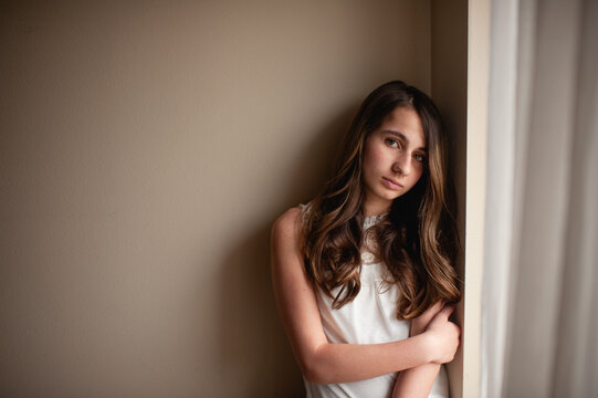 Teen girl leans on wall with neutral expression by window at home