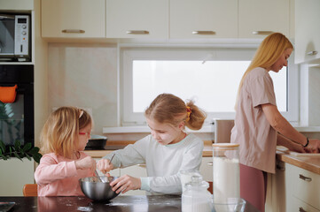 Kids stirring thick dough with wooden spoons at the table while their mother prepares food on the stove creating a lively yet warm family kitchen atmosphere