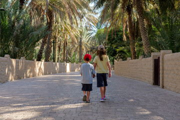 Two kids walk through Al Ain Oasis, UAE, a popular tourist spot. The oasis offers shade and a cool escape from the heat, making it a perfect place for a relaxing walk in nature