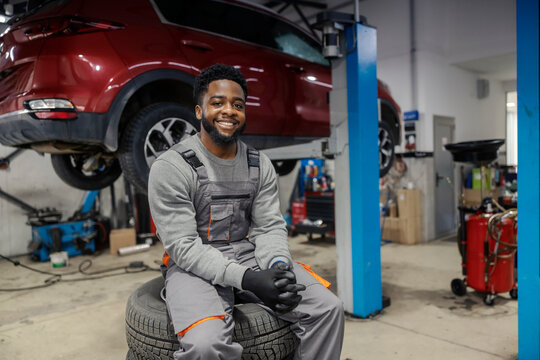 Portrait of happy interracial repairman sitting on tires at auto mechanic workshop and smiling at camera.